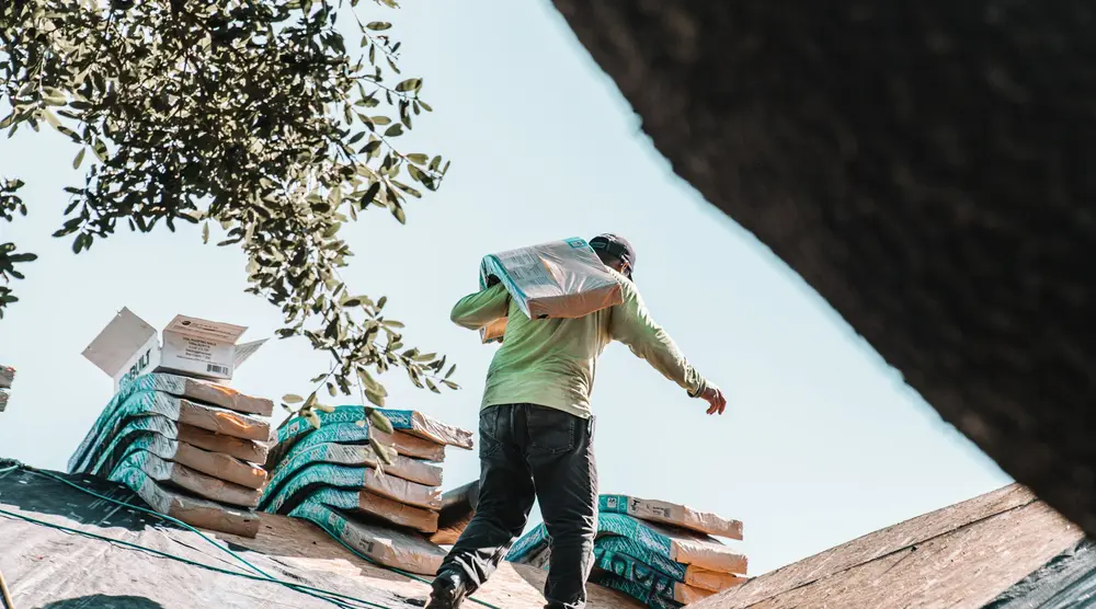 A worker carrying a bundle of shingles across a roof under construction.