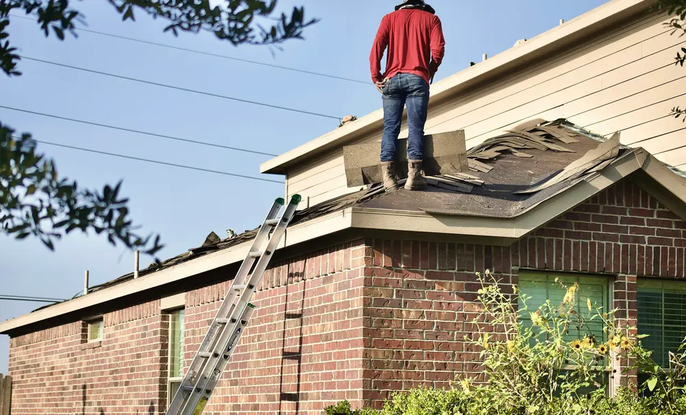 A worker standing on the roof of a brick house beside a ladder.