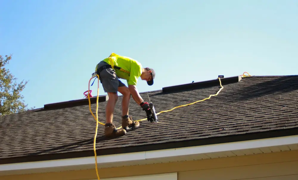 A roofer checking a shingle roof beneath a wide blue sky.